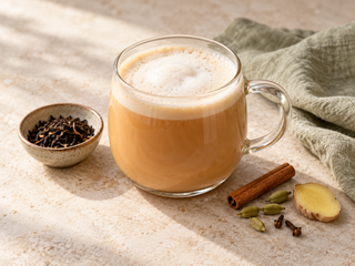 Warmer Chai Latte mit Milchschaum in einer Glastasse, umgeben von Zimt, Kardamom, Ingwer und Schwarztee.