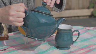 Tee wird aus einer blauen Porzellan Kanne in eine blaue Porzellan Tasse gegossen. Im Hintergrund sind zwei scheiben Toast und eine Flasche Honig zu sehen. 