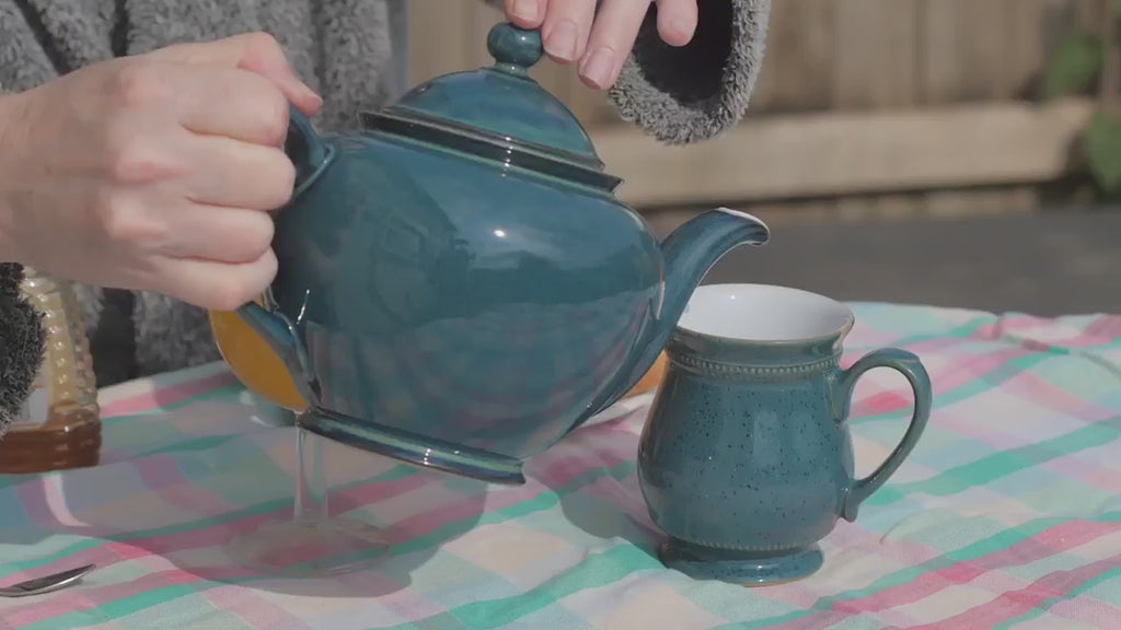 Tee wird aus einer blauen Porzellan Kanne in eine blaue Porzellan Tasse gegossen. Im Hintergrund sind zwei scheiben Toast und eine Flasche Honig zu sehen. 